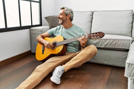 Photo for Middle age grey-haired man playing classical guitar sitting on floor at home - Royalty Free Image