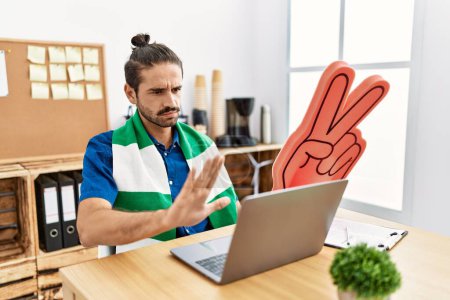 Photo for Young hispanic man watching football on laptop cheering game at the office with open hand doing stop sign with serious and confident expression, defense gesture - Royalty Free Image