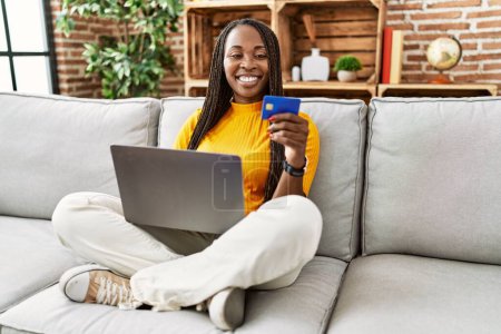 Foto de Mujer afroamericana usando laptop y tarjeta de crédito sentada en sofá en casa - Imagen libre de derechos