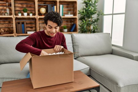 Photo for Young hispanic man unboxing cardboard box sitting on the sofa at home. - Royalty Free Image