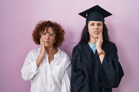 Foto de Hispanic mother and daughter wearing graduation cap and ceremony robe touching mouth with hand with painful expression because of toothache or dental illness on teeth. dentist - Imagen libre de derechos