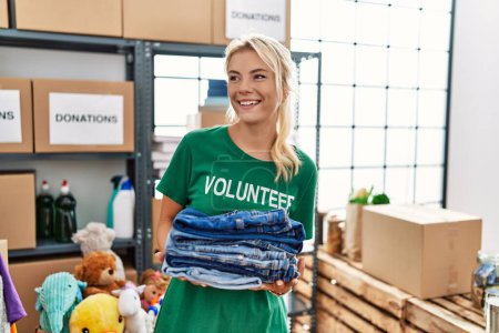 Téléchargez les photos : Jeune fille volontaire blonde souriant heureux tenant pile de jeans pliés au centre de charité. - en image libre de droit