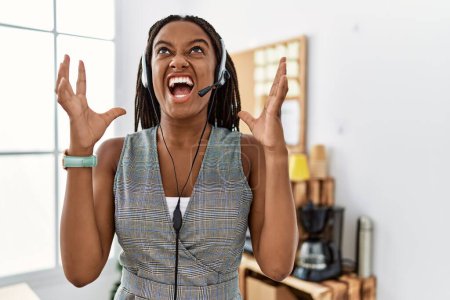 Téléchargez les photos : Jeune femme afro-américaine travaillant au bureau portant casque opérateur fou et fou crier et crier avec une expression agressive et les bras levés. concept de frustration. - en image libre de droit
