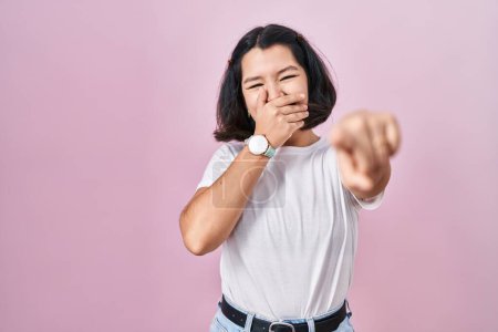 Téléchargez les photos : Jeune femme hispanique portant un t-shirt blanc décontracté sur fond rose se moquant de vous, pointant du doigt l'appareil photo avec la main sur la bouche, expression de honte - en image libre de droit