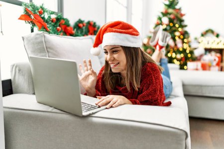 Téléchargez les photos : Jeune fille hispanique ayant appel vidéo couché sur le canapé par arbre de Noël à la maison. - en image libre de droit