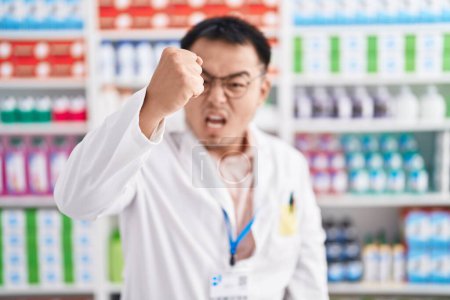 Téléchargez les photos : Jeune homme chinois travaillant à la pharmacie pharmacie en colère et fou élever le poing frustré et furieux tout en criant avec colère. rage et concept agressif. - en image libre de droit