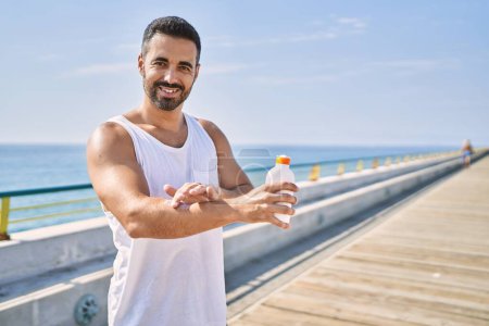 Téléchargez les photos : Homme de sport hispanique portant un style d'entraînement appliquant de la crème solaire sur le bras à l'extérieur par une journée ensoleillée - en image libre de droit