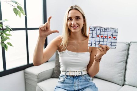 Foto de Young blonde woman holding heart calendar smiling and confident gesturing with hand doing small size sign with fingers looking and the camera. measure concept. - Imagen libre de derechos