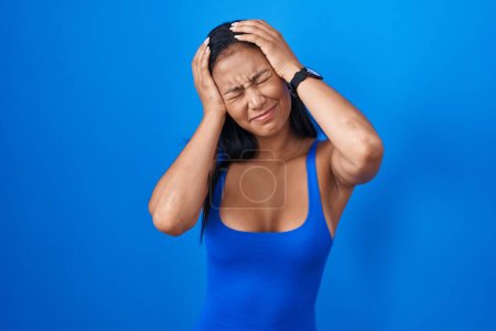 Foto de Hispanic woman standing over blue background suffering from headache desperate and stressed because pain and migraine. hands on head. - Imagen libre de derechos