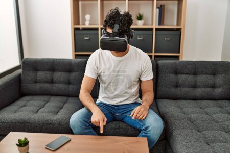Photo for Young hispanic man playing with virtual reality glasses sitting on the sofa at home. - Royalty Free Image