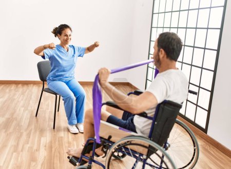 Foto de Hombre y mujer de mediana edad que tienen sesión de rehabilitación con banda elástica sentados en silla de ruedas en la clínica de fisioterapia - Imagen libre de derechos