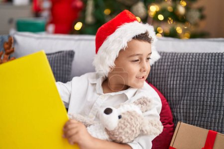 Foto de Adorable niño hispano leyendo libro sentado en el sofá junto al árbol de Navidad en casa - Imagen libre de derechos