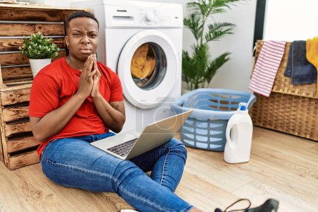 Téléchargez les photos : Jeune homme africain faire la lessive et en utilisant l'ordinateur mendier et prier avec les mains ainsi que l'expression de l'espoir sur le visage très émotionnel et inquiet. mendier. - en image libre de droit