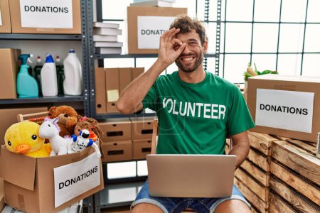 Téléchargez les photos : Young handsome man wearing volunteer t shirt using laptop doing ok gesture with hand smiling, eye looking through fingers with happy face. - en image libre de droit