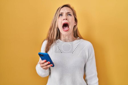 Photo for Young blonde woman using smartphone typing message angry and mad screaming frustrated and furious, shouting with anger. rage and aggressive concept. - Royalty Free Image