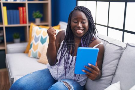 Téléchargez les photos : Jeune femme africaine utilisant touchpad assis sur le canapé criant fier, célébrant la victoire et le succès très excité avec le bras levé - en image libre de droit