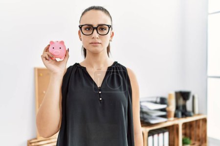 Photo for Young brunette woman holding piggy bank at the office thinking attitude and sober expression looking self confident - Royalty Free Image