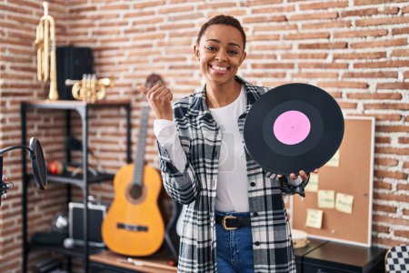 Foto de Hermosa mujer afroamericana sosteniendo un disco de vinilo en un estudio de música apuntando con el pulgar hacia un lado sonriendo feliz con la boca abierta - Imagen libre de derechos