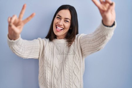 Téléchargez les photos : Jeune femme brune debout sur fond bleu souriant avec la langue montrant les doigts des deux mains faisant signe de victoire. numéro deux. - en image libre de droit
