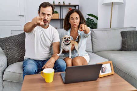 Téléchargez les photos : Couple d'âge moyen hispanique assis sur le canapé à l'aide d'un ordinateur portable pointant du doigt vers la caméra et à vous, geste confiant regardant sérieux - en image libre de droit