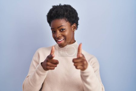 Foto de Mujer afroamericana de pie sobre fondo azul señalando los dedos a la cámara con la cara feliz y divertida. buena energía y vibraciones. - Imagen libre de derechos