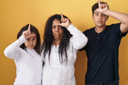 Téléchargez les photos : Family of mother, daughter and son standing over yellow background making fun of people with fingers on forehead doing loser gesture mocking and insulting. - en image libre de droit