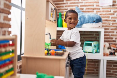 Téléchargez les photos : Africain américain garçon souriant confiant jouer avec la cuisine de jeu à la maternelle - en image libre de droit