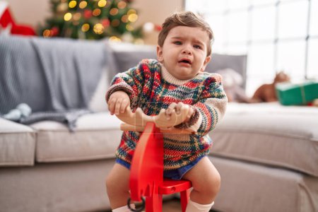 Foto de Adorable niño hispano llorando y jugando con renos meciéndose en el árbol de Navidad en casa - Imagen libre de derechos