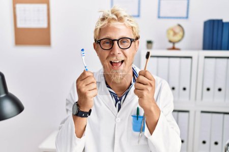 Foto de Joven dentista rubio trabajando en una clínica de dentistas sosteniendo cepillos de dientes sonriendo y riendo en voz alta porque gracioso chiste loco. - Imagen libre de derechos