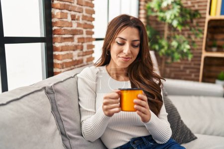 Foto de Mujer hispana joven bebiendo café sentada en el sofá en casa - Imagen libre de derechos