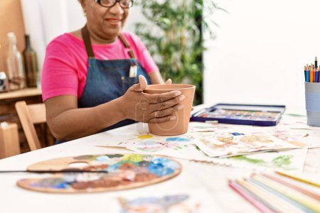 Téléchargez les photos : Femme afro-américaine senior souriant confiant faire de la céramique d'argile au studio d'art - en image libre de droit
