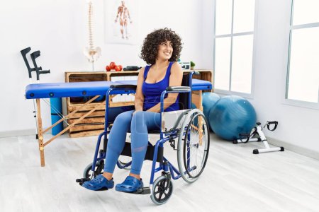 Foto de Young middle eastern woman sitting on wheelchair at physiotherapy clinic looking away to side with smile on face, natural expression. laughing confident. - Imagen libre de derechos