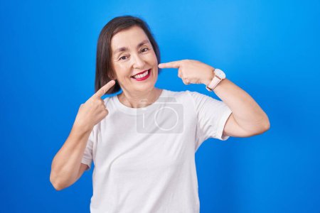 Photo for Middle age hispanic woman standing over blue background smiling cheerful showing and pointing with fingers teeth and mouth. dental health concept. - Royalty Free Image