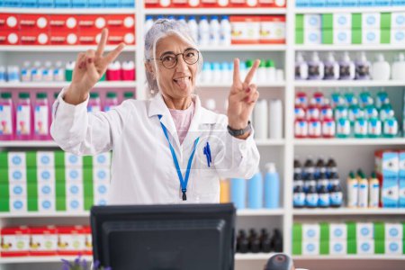 Photo for Middle age woman with tattoos working at pharmacy drugstore smiling with tongue out showing fingers of both hands doing victory sign. number two. - Royalty Free Image