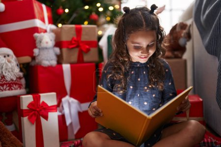 Foto de Adorable chica hispana leyendo libro sentado en el suelo por árbol de Navidad en casa - Imagen libre de derechos