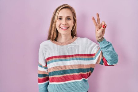 Foto de Mujer rubia joven de pie sobre fondo rosa mostrando y señalando hacia arriba con los dedos número dos mientras sonríe confiado y feliz. - Imagen libre de derechos
