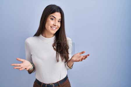 Téléchargez les photos : Jeune femme brune debout sur fond bleu souriant gai avec les bras ouverts comme accueil amical, salutations positives et confiantes - en image libre de droit