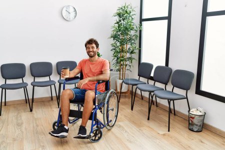 Foto de Joven hispano bebiendo café sentado en silla de ruedas en la sala de espera de la clínica. - Imagen libre de derechos