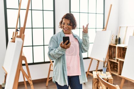 Foto de Mujer artista hispana de mediana edad sonriendo feliz teniendo videollamada usando teléfono inteligente en el estudio de arte. - Imagen libre de derechos