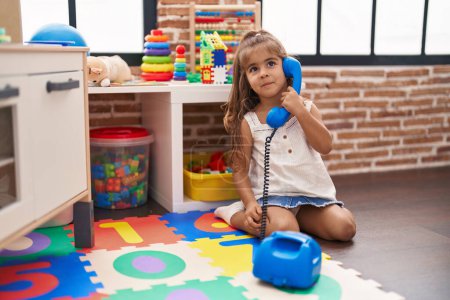 Téléchargez les photos : Adorable fille hispanique jouant jouet téléphonique assis sur le sol à la maternelle - en image libre de droit