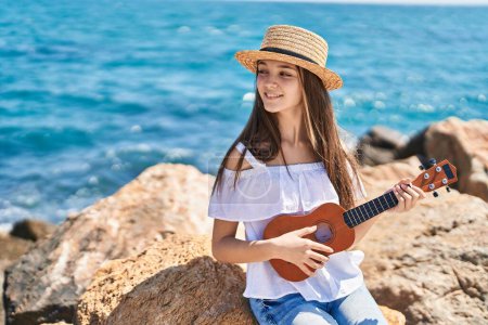 Téléchargez les photos : Adorable fille touristique souriant confiant jouer ukulele au bord de la mer - en image libre de droit