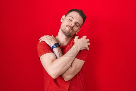 Photo for Young hispanic man standing over red background hugging oneself happy and positive, smiling confident. self love and self care - Royalty Free Image