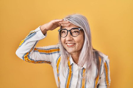 Photo for Middle age woman with grey hair standing over yellow background wearing glasses very happy and smiling looking far away with hand over head. searching concept. - Royalty Free Image