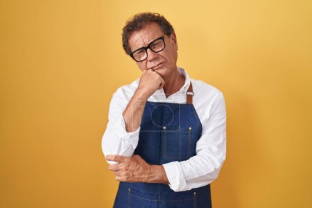 Middle age hispanic man wearing professional cook apron with hand on chin thinking about question, pensive expression. smiling with thoughtful face. doubt concept. 