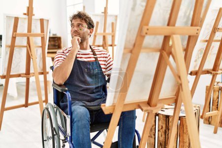 Foto de Young handsome man with beard at art studio sitting on wheelchair serious face thinking about question with hand on chin, thoughtful about confusing idea - Imagen libre de derechos