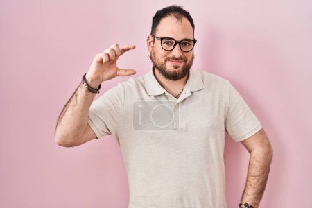 Téléchargez les photos : Plus size hispanic man with beard standing over pink background smiling and confident gesturing with hand doing small size sign with fingers looking and the camera. measure concept. - en image libre de droit