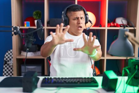 Téléchargez les photos : Jeune homme hispanique jouant à des jeux vidéo effrayé et terrifié par l'expression de la peur arrêter geste avec les mains, criant en état de choc. concept de panique. - en image libre de droit