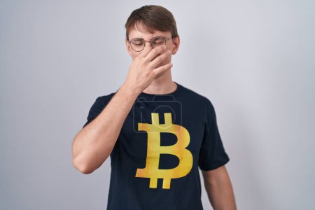 Foto de Caucasian blond man wearing bitcoin t shirt smelling something stinky and disgusting, intolerable smell, holding breath with fingers on nose. bad smell - Imagen libre de derechos