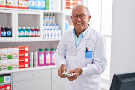 Foto de Hombre mayor de pelo gris farmacéutico sonriendo confiado sosteniendo pastillas tabletas en la farmacia - Imagen libre de derechos
