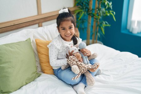 Foto de Adorable hispanic girl holding dolls sitting on bed at bedroom - Imagen libre de derechos